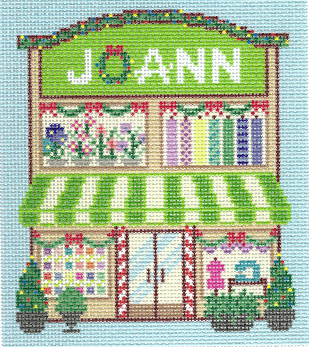Haus of Toots needlepoint canvas design of a storefront with the word "JOANN" in white letters on a green sign above windows displaying colorful fabrics and sewing notions. The awning is green and white striped, and the scene is set against a light blue background with Christmas trees out front.