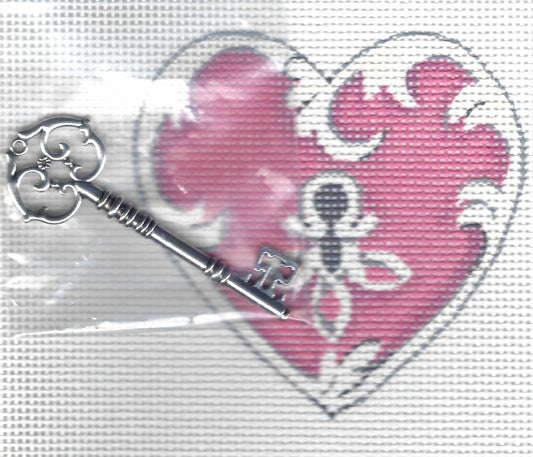 A silver-colored metal key with an ornate decorative bow, shown in front of a needlepoint canvas featuring a pink and white heart-shaped pattern with a keyhole.