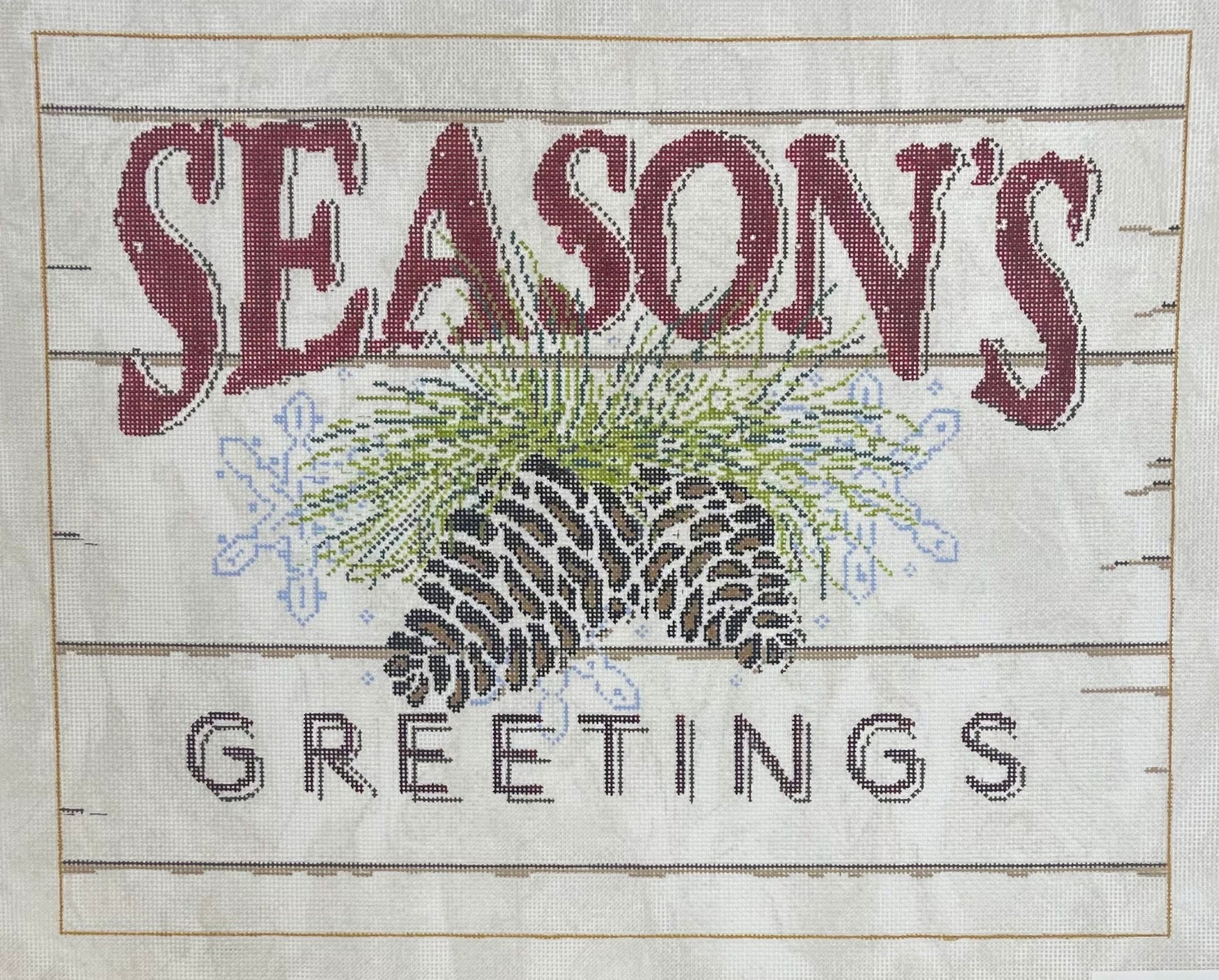 Sandra Gilmore needlepoint canvas of a sign with the words “SEASON'S GREETINGS” stitched on a light background with horizontal wood-plank lines. The center features pinecones, green pine needles, and small blue snowflakes.