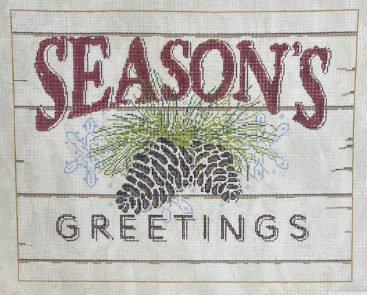 Sandra Gilmore needlepoint canvas of a sign with the words “SEASON'S GREETINGS” stitched on a light background with horizontal wood-plank lines. The center features pinecones, green pine needles, and small blue snowflakes.