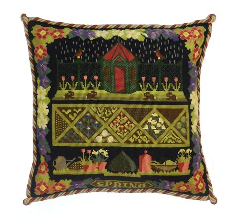 Elizabeth Bradley square cross stitch tapestry kit of pillow featuring a spring garden scene with a small red-roofed cottage, flowers, and geometric patterns on a black background, finished with a multicolored twisted rope border.