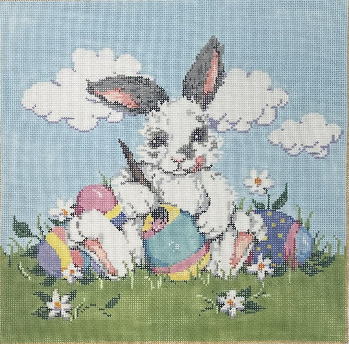 Sandra Gilmore needlepoint canvas of a bunny sitting in a field with four colorful Easter eggs with a light blue sky background featuring clouds. 