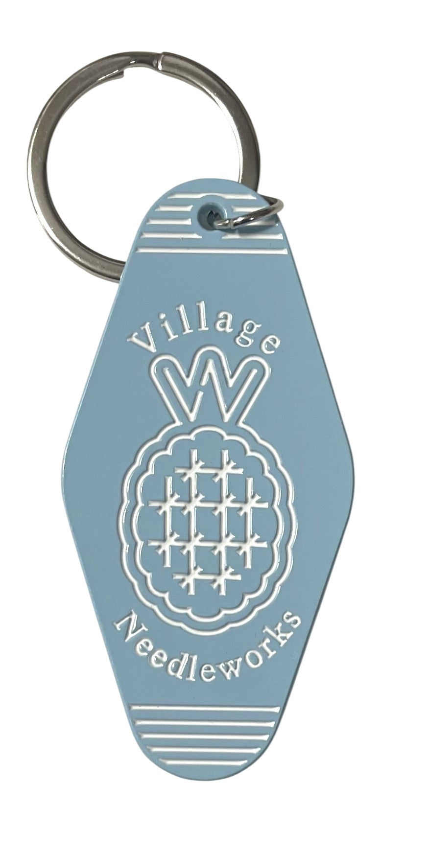 A light blue keychain with a silver metal split ring, featuring a white engraved pineapple design and the text 'Village Needleworks'.