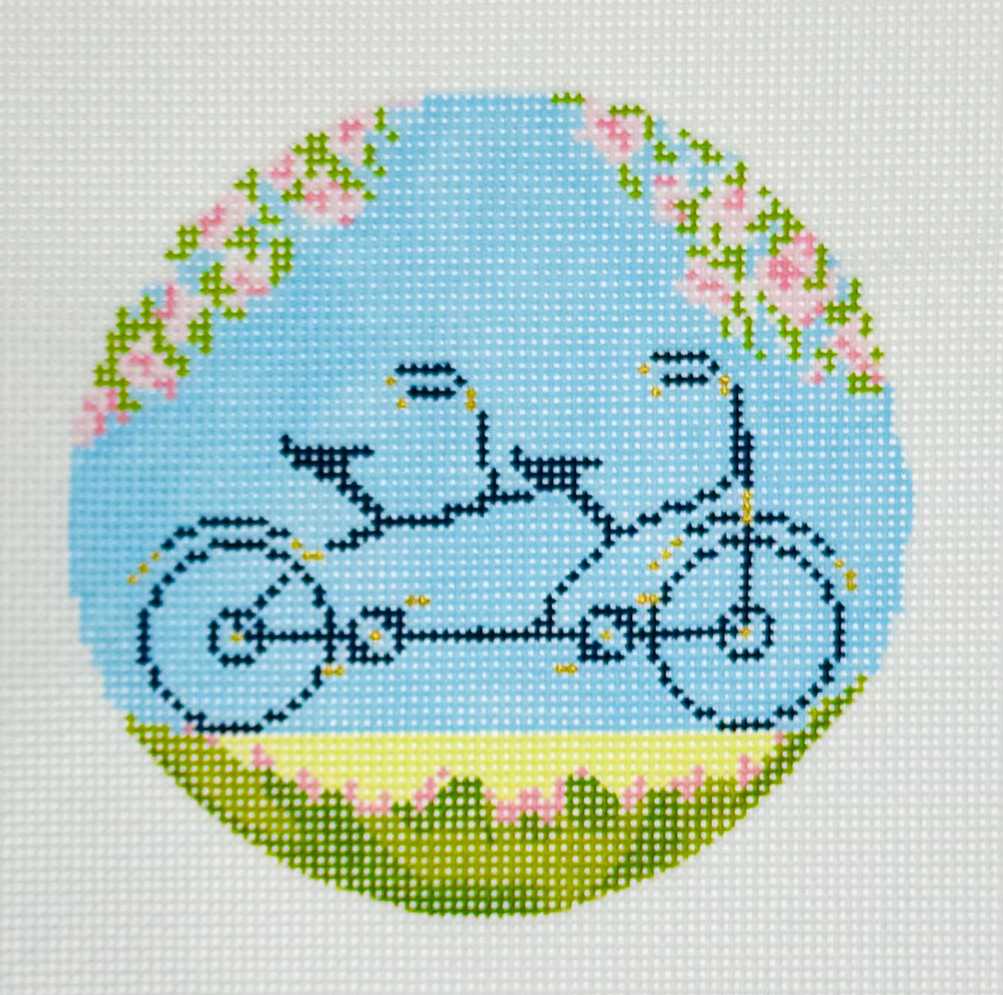 The Plum Stitchery round needlepoint canvas of a tandem bicycle built for two with flowers and a light blue background.