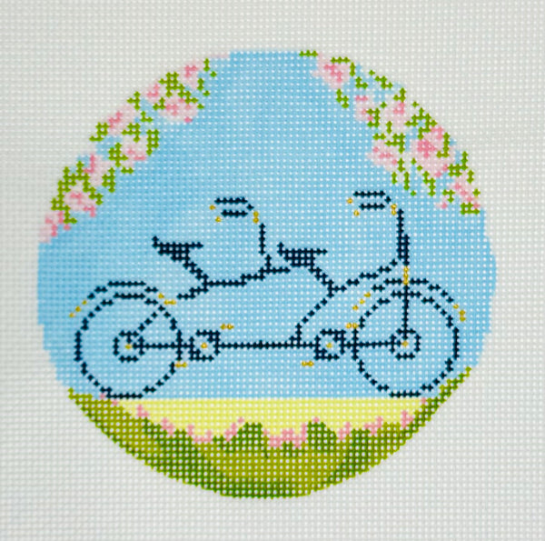 The Plum Stitchery round needlepoint canvas of a tandem bicycle built for two with flowers and a light blue background.
