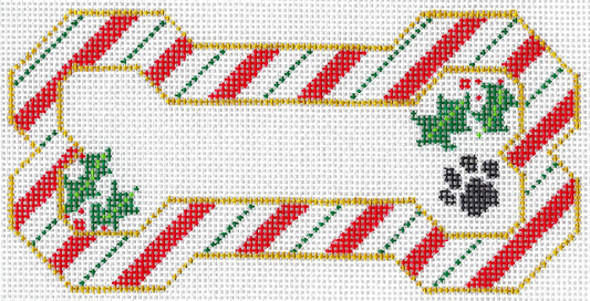 Danji Designs needlepoint ornament canvas featuring a bone-shaped border with red and white diagonal stripes, green holly leaves, red berries, and a black paw print.