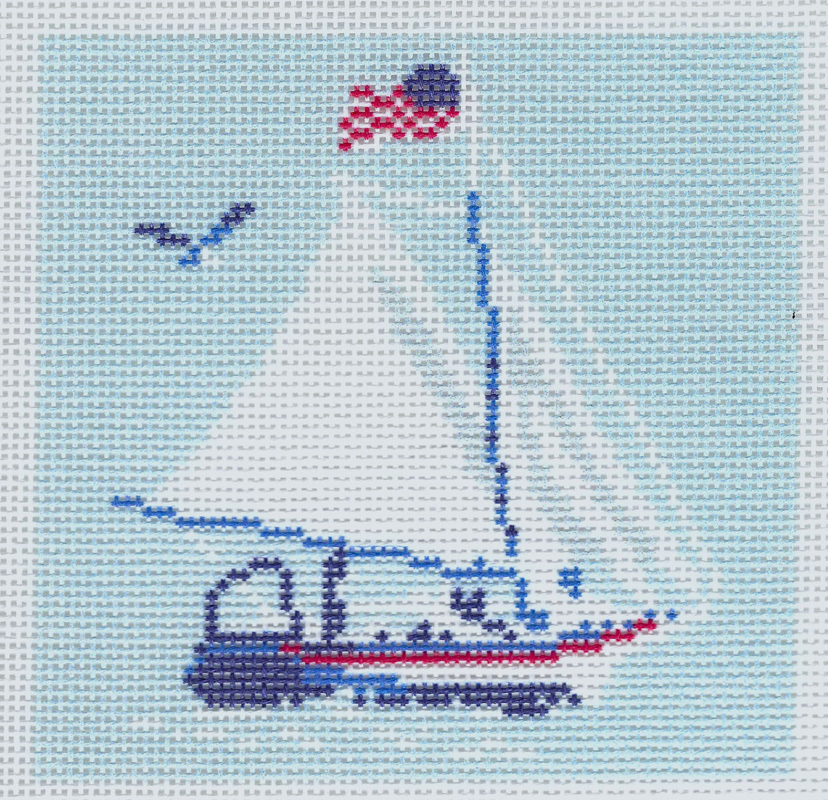 Needlepoint To Go needlepoint canvas of a sailboat with a white sail, a blue hull, a red stripe, and an American flag on the mast, set against a light blue background with a small blue bird.