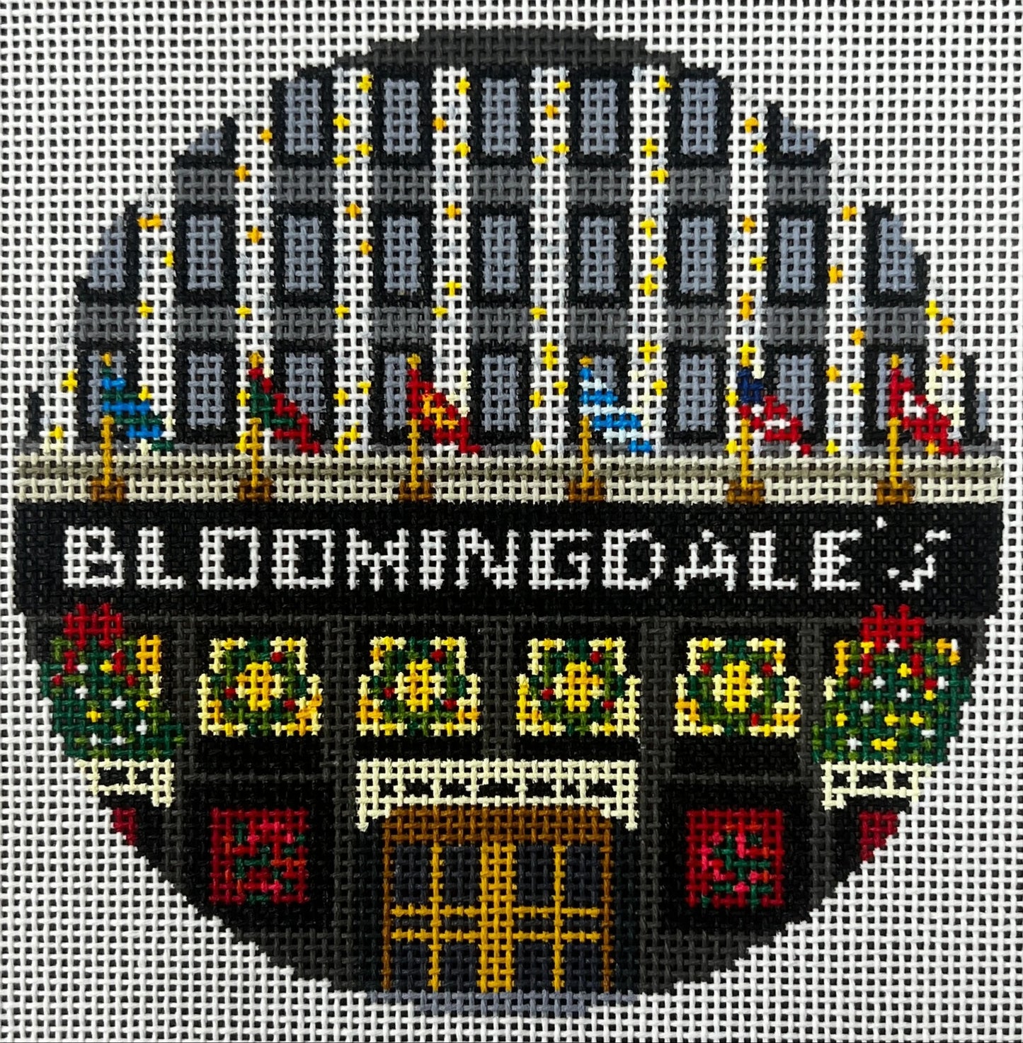 Gingham Stitchery circular needlepoint canvas of the words 'BERGDORT GODMAN' in a white sign above an arched doorway with yellow and orange window light, flanked by floral panels and gray side sections on a light background.