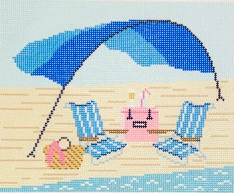 Needlepoint To Go needlepoint canvas showing a blue umbrella on a beach with two blue lounge chairs, a pink cooler, a small pink umbrella, and a beach bag on a sandy shore with light blue water.